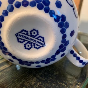 May include: White ceramic mug with a blue and white geometric pattern. The mug has a handle and the bottom of the mug has the word "Anthropologie" printed on it.