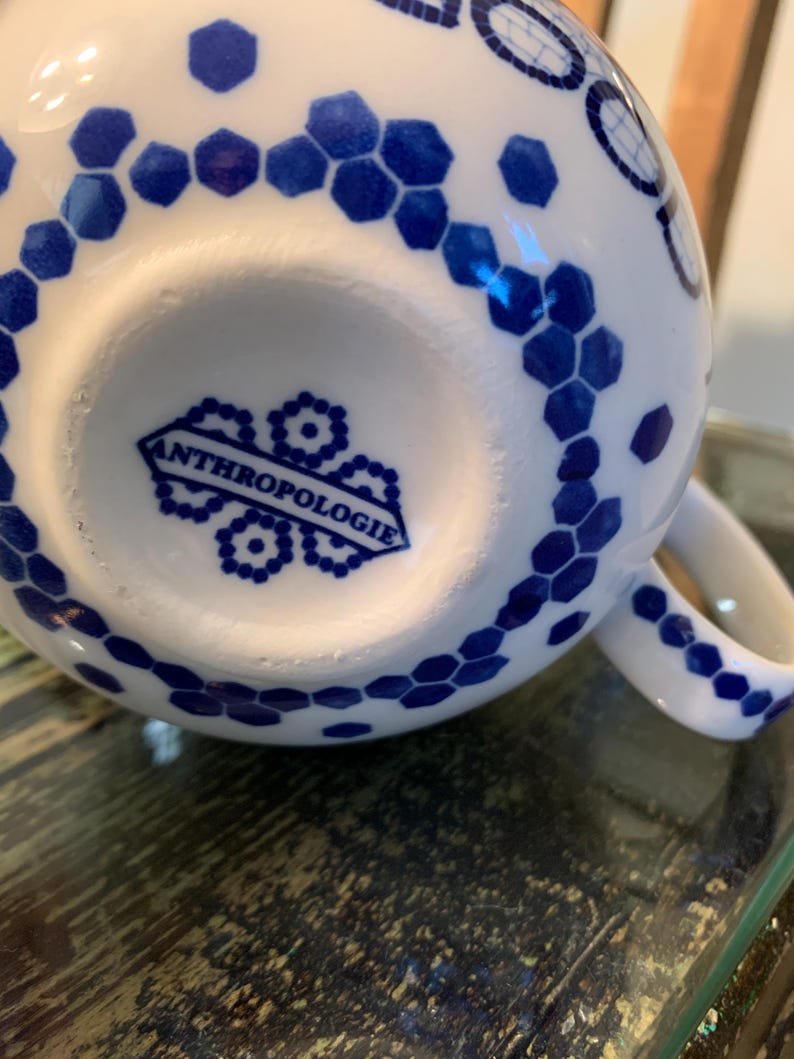May include: White ceramic mug with a blue and white geometric pattern. The mug has a handle and the bottom of the mug has the word "Anthropologie" printed on it.
