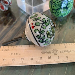May include: A white ceramic knob with a green and red floral design. The knob is attached to a metal screw and is about 2 inches in diameter.
