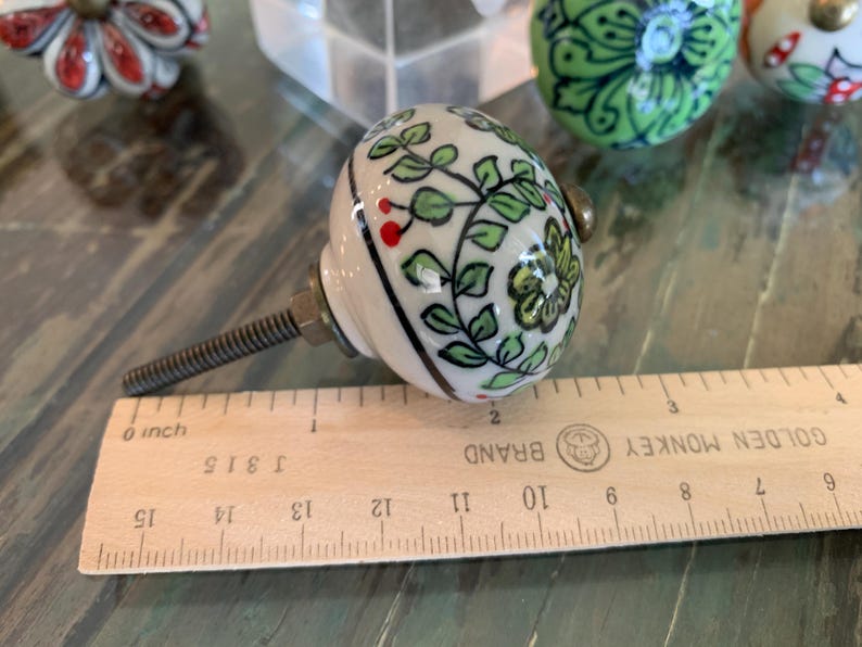May include: A white ceramic knob with a green and red floral design. The knob is attached to a metal screw and is about 2 inches in diameter.