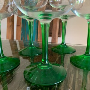 May include: A collection of clear glass goblets with vibrant green stems and bases. The glasses are arranged on a reflective surface, showcasing their elegant design. The green color adds a pop of color to the glassware.