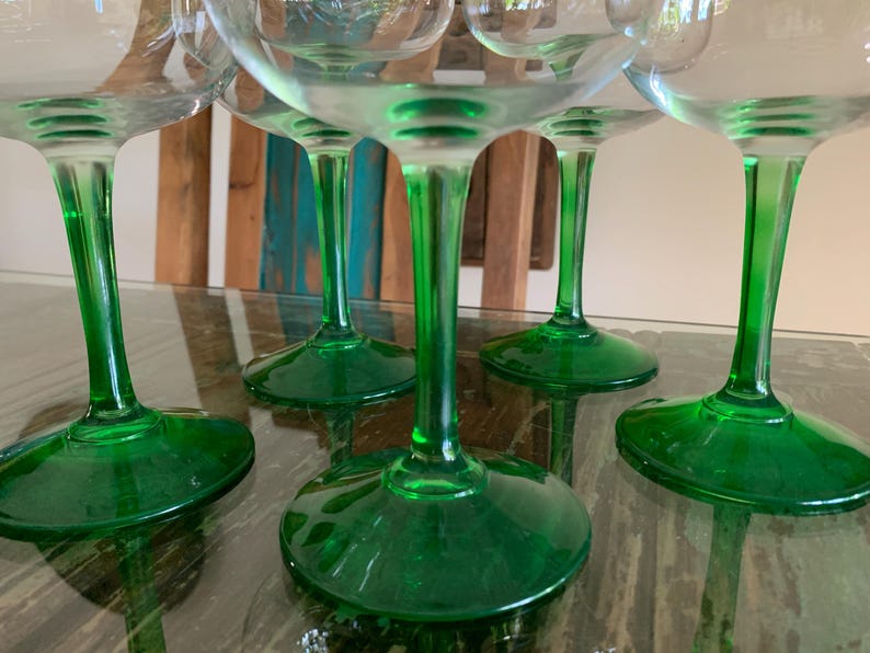 May include: A collection of clear glass goblets with vibrant green stems and bases. The glasses are arranged on a reflective surface, showcasing their elegant design. The green color adds a pop of color to the glassware.