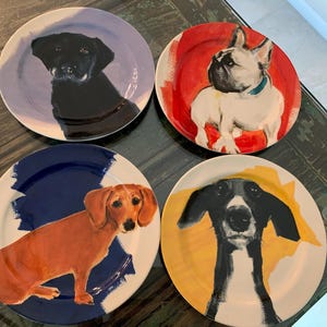 May include: Four ceramic plates with dog portraits. Each plate features a different dog breed: a black Labrador, a French Bulldog, a Dachshund, and a black and white dog. The plates have colorful abstract backgrounds.