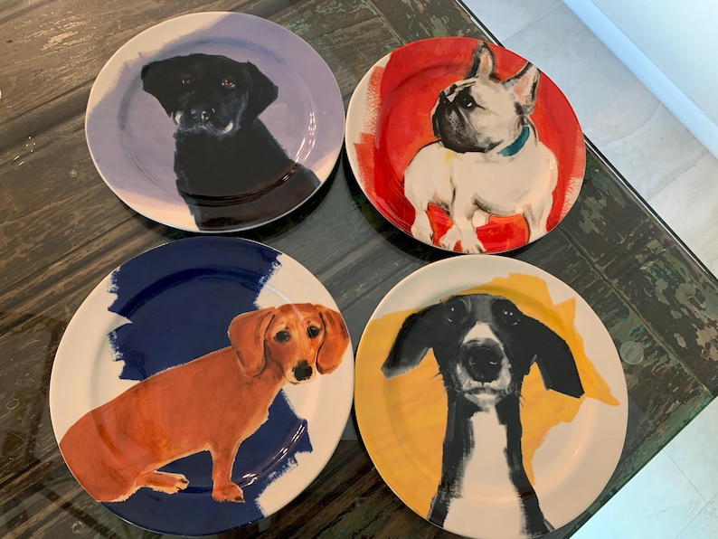 May include: Four ceramic plates with dog portraits. Each plate features a different dog breed: a black Labrador, a French Bulldog, a Dachshund, and a black and white dog. The plates have colorful abstract backgrounds.