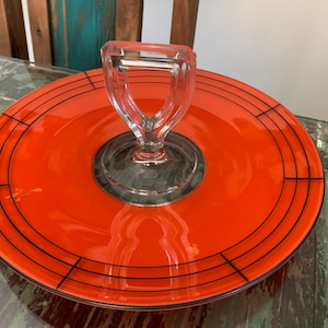 May include: A vibrant orange glass serving tray with a clear glass handle. The tray features black linear accents and a circular design. This decorative piece is ideal for serving appetizers or desserts.