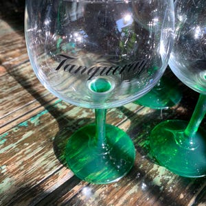 May include: Clear glass gin glasses with green stems and bases. The word "Tanqueray" is printed in black on the front of the glass. The glasses are on a weathered wooden surface.