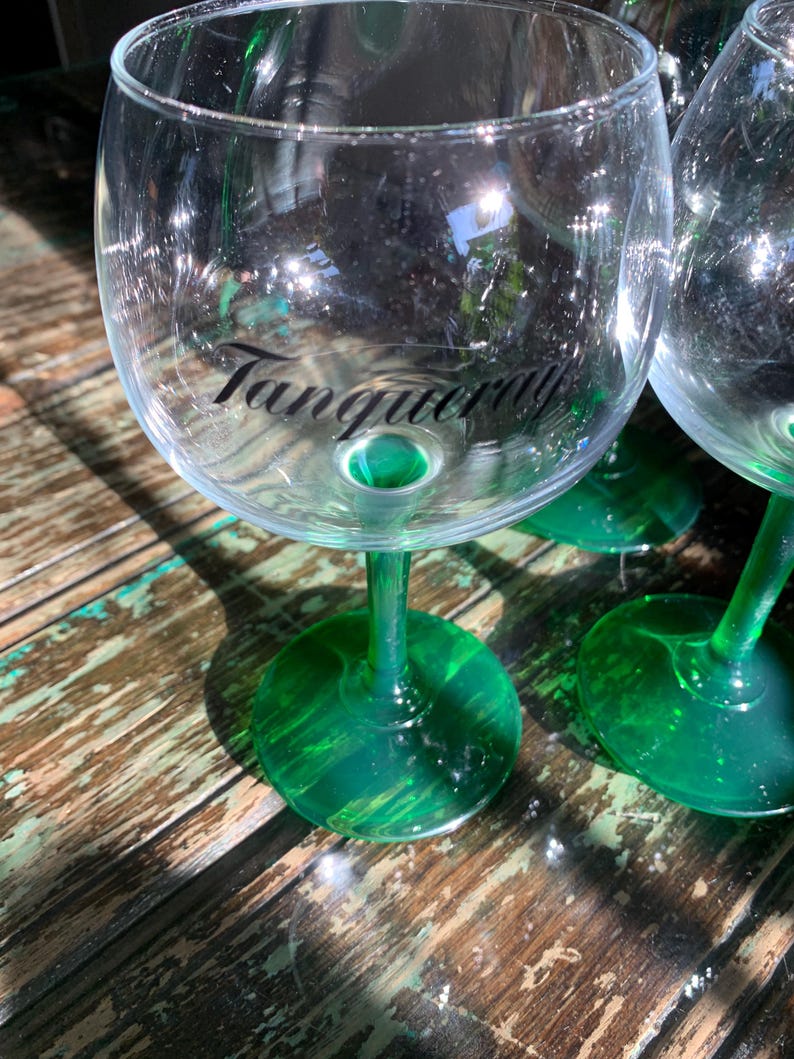 May include: Clear glass gin glasses with green stems and bases. The word "Tanqueray" is printed in black on the front of the glass. The glasses are on a weathered wooden surface.