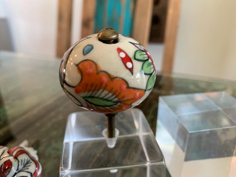 May include: A white ceramic knob with a floral design in shades of orange, green, and blue. The knob has a brass screw and is mounted on a clear acrylic base.