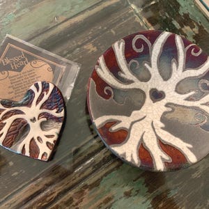 May include: A heart-shaped ceramic piece and a circular ceramic dish, both featuring a white tree design with a small heart at the center against a crackled, multi-colored background of deep red, blue, and purple tones, with a small card labeled Blessed Heart.