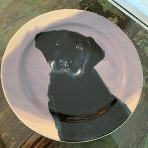 May include: A round ceramic plate featuring a black Labrador Retriever portrait. The dog's head and shoulders are depicted against a lavender-colored background. The plate has a white rim and is suitable for serving food.