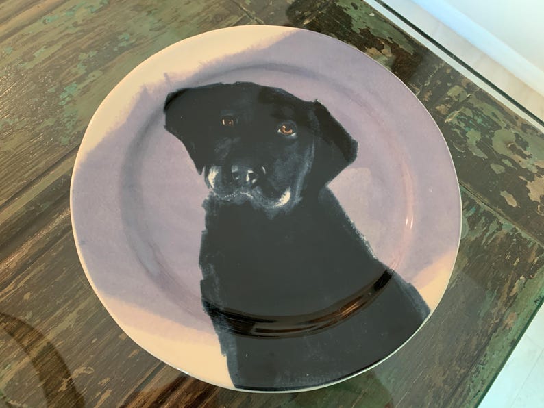 May include: A round ceramic plate featuring a black Labrador Retriever portrait. The dog's head and shoulders are depicted against a lavender-colored background. The plate has a white rim and is suitable for serving food.