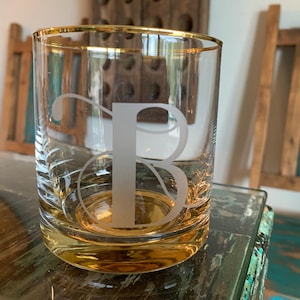 May include: A clear glass tumbler with a gold rim and base, featuring a frosted letter "B". The glass is set on a reflective surface, with a blurred background of wooden furniture. This personalized glass is ideal for beverages.