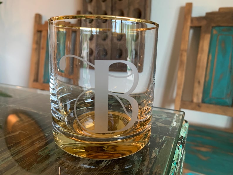 May include: A clear glass tumbler with a gold rim and base, featuring a frosted letter "B". The glass is set on a reflective surface, with a blurred background of wooden furniture. This personalized glass is ideal for beverages.