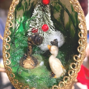May include: A miniature Christmas scene inside a decorated eggshell ornament. A small white duck stands near a tiny Christmas tree with red ornaments and a black lamppost.