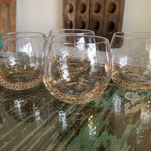 May include: Four clear, stemless wine glasses with a gold polka dot pattern around the base. The glasses are sitting on a green and brown surface. A wooden wine rack is in the background.