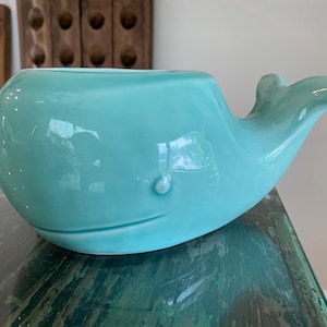 May include: A glossy, light turquoise ceramic whale-shaped bowl or planter. The object features a smooth, rounded body, a small raised eye detail, and a curved tail fin extending upward on the right side. The surface has a reflective finish.