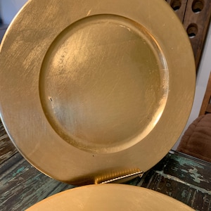 May include: A round charger plate with a metallic gold finish and a smooth, reflective surface. The plate features a wide, flat rim and a slightly recessed center, displaying subtle brush-like textures across the circular surface.