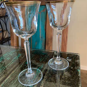 May include: Two clear glass wine glasses with gold rims on a wooden table. The glasses are empty and have a classic design.