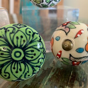 May include: Two ceramic knobs with floral designs. One knob is green with black floral details, and the other is white with multi-colored floral details.