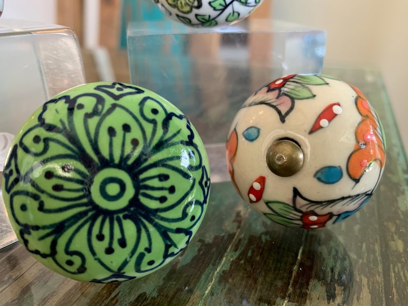 May include: Two ceramic knobs with floral designs. One knob is green with black floral details, and the other is white with multi-colored floral details.