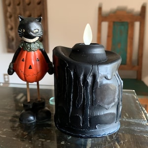 May include: A black Halloween candle with a dripping wax design and a flickering LED flame. Beside it is a black cat figurine with a pumpkin body and wooden legs. The cat has a smiling face and is wearing a green collar.