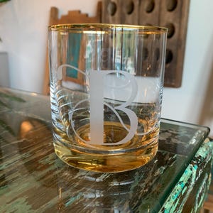 May include: A clear glass tumbler with a gold rim and a frosted letter "B" etched on the front. The glass sits on a distressed, multi-colored surface. The tumbler is designed for beverages.