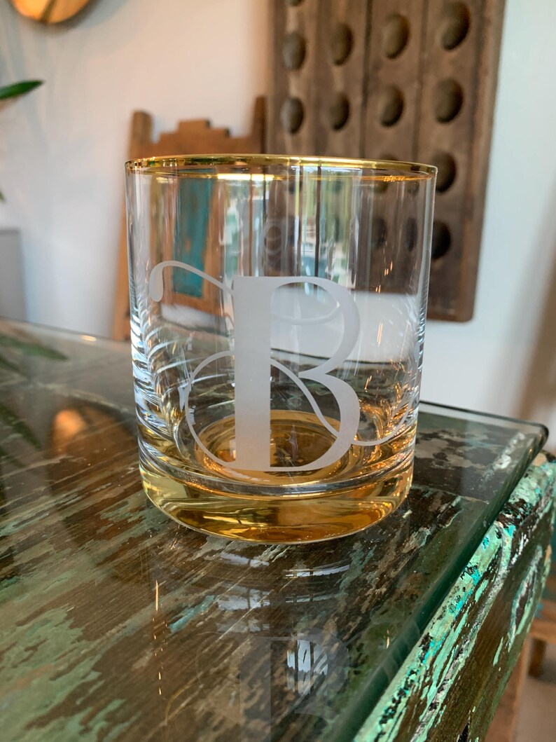 May include: A clear glass tumbler with a gold rim and a frosted letter "B" etched on the front. The glass sits on a distressed, multi-colored surface. The tumbler is designed for beverages.