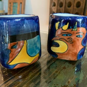 May include: Two hand-painted ceramic cups with a dark blue glaze. One cup features a colorful abstract face design, while the other displays a cartoonish orange creature with yellow horns and blue eyes. The cups are on a wooden table.
