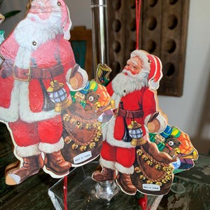 May include: Cardboard Santa Claus Christmas ornaments. The ornaments are red, white, and brown, with a detailed illustration of Santa Claus holding a lantern and a sack of toys. The ornaments are hanging from red strings.