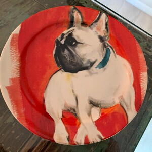 May include: A round decorative plate featuring a French Bulldog illustration. The dog is white with black accents and a blue collar, set against a red and white background. The plate is likely ceramic.