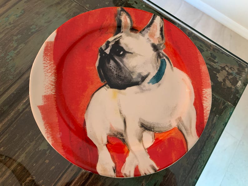 May include: A round decorative plate featuring a French Bulldog illustration. The dog is white with black accents and a blue collar, set against a red and white background. The plate is likely ceramic.