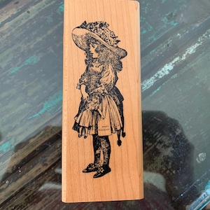 May include: A rectangular wooden stamp featuring a detailed black and white illustration of a young girl in a large hat and dress, holding a doll. The girl is standing, and the stamp is made of light-colored wood.