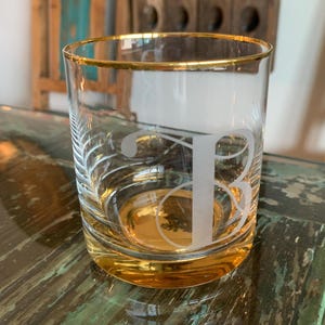 May include: A clear glass tumbler with a gold rim and base. The glass features an etched letter "B" and a decorative leaf design. The tumbler is partially filled with a golden liquid.