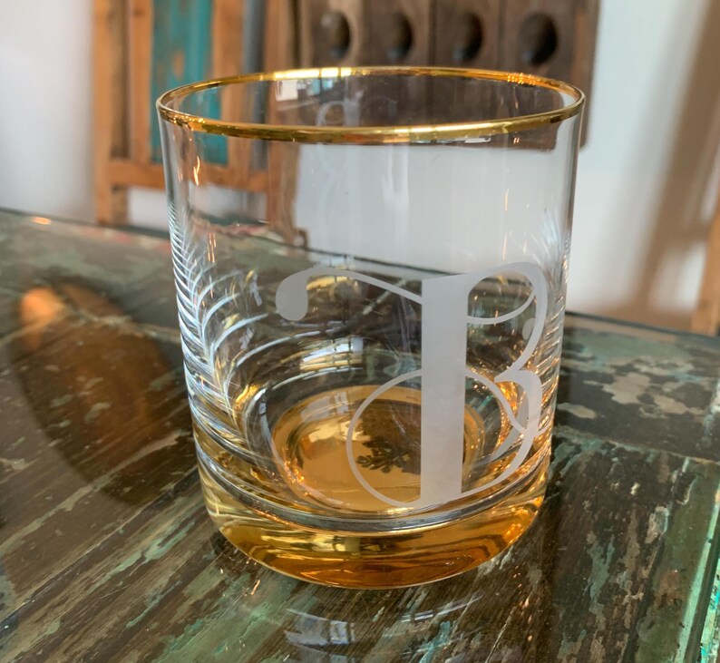 May include: A clear glass tumbler with a gold rim and base. The glass features an etched letter "B" and a decorative leaf design. The tumbler is partially filled with a golden liquid.