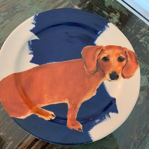 May include: A round ceramic plate featuring a dachshund dog illustration. The dog is depicted in shades of brown, set against a blue and white background. The plate's design has a painted aesthetic, suitable for decorative or functional use.