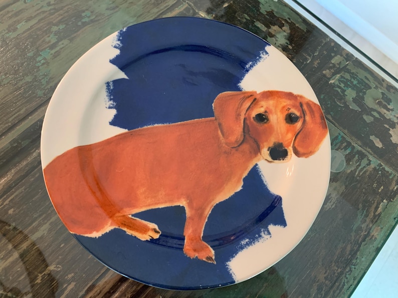 May include: A round ceramic plate featuring a dachshund dog illustration. The dog is depicted in shades of brown, set against a blue and white background. The plate's design has a painted aesthetic, suitable for decorative or functional use.