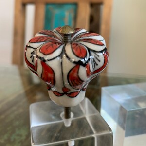 May include: A white ceramic cabinet knob with a red and black floral design. The knob is mounted on a clear acrylic base.