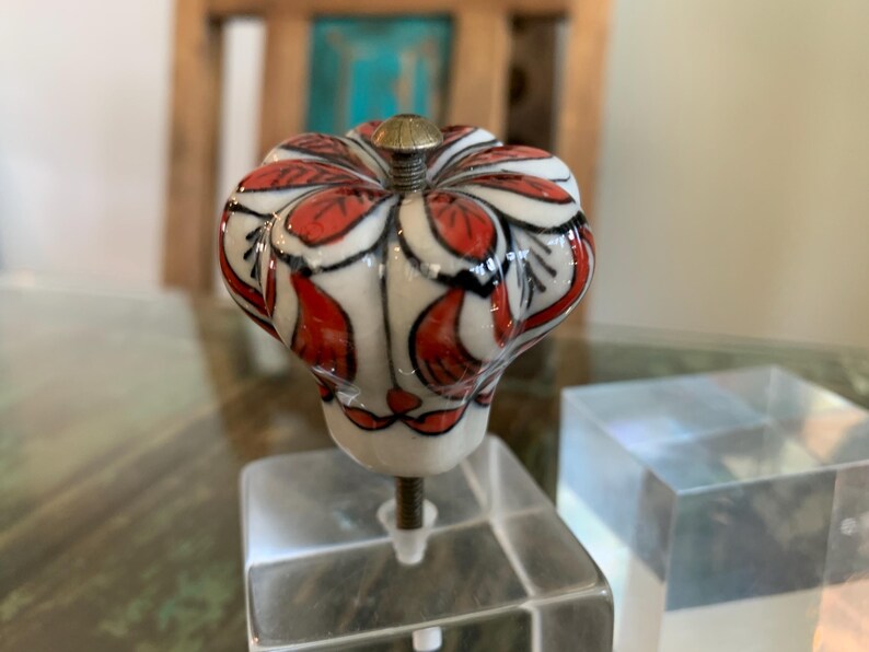 May include: A white ceramic cabinet knob with a red and black floral design. The knob is mounted on a clear acrylic base.