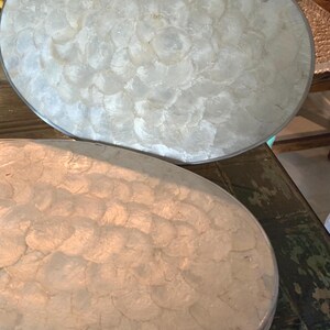 May include: An oval-shaped box with a hinged lid, featuring a surface covered in overlapping, translucent white mother-of-pearl shell discs. The container has a thin, metallic silver-toned rim outlining both the lid and the base structure.