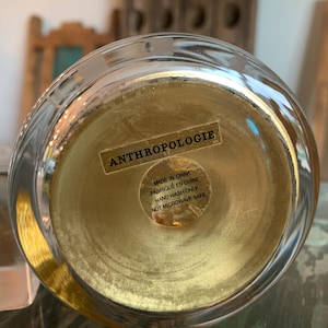 May include: A clear glass bowl with a gold-colored interior. The bottom of the bowl has a sticker that reads "ANTHROPOLOGIE", "MADE IN CHINA", "FABRIQU&Eacute; EN CHINE", "HAND WASH ONLY", and "NOT MICROWAVE SAFE".