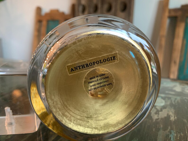 May include: A clear glass bowl with a gold-colored interior. The bottom of the bowl has a sticker that reads "ANTHROPOLOGIE", "MADE IN CHINA", "FABRIQU&Eacute; EN CHINE", "HAND WASH ONLY", and "NOT MICROWAVE SAFE".