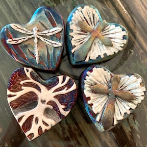 May include: Four heart-shaped ceramic tiles. One tile features a dragonfly, another a tree, and two others have a cross design. The tiles have a mix of blue, red, and white colors, with a glossy finish.