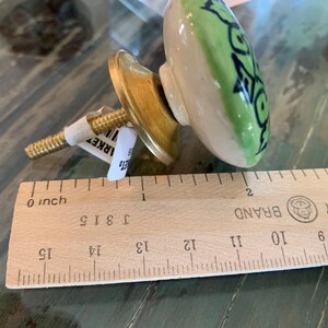 May include: A green and white ceramic knob with a black floral design. The knob is attached to a gold metal base with a screw. The knob is measured against a wooden ruler.