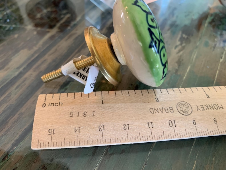 May include: A green and white ceramic knob with a black floral design. The knob is attached to a gold metal base with a screw. The knob is measured against a wooden ruler.