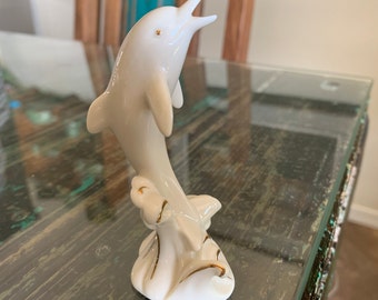 May include: White porcelain dolphin figurine with gold accents, leaping out of the water. The dolphin is positioned on a base with stylized waves and leaves.