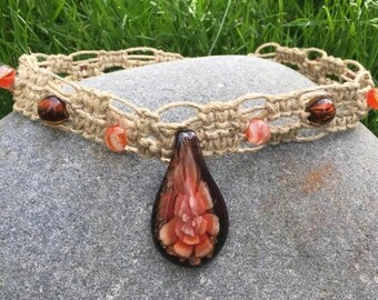 May include: A macrame choker necklace with a teardrop pendant. The necklace is made of tan-colored hemp cord and adorned with orange and brown beads. The pendant features a floral design in shades of orange and brown.