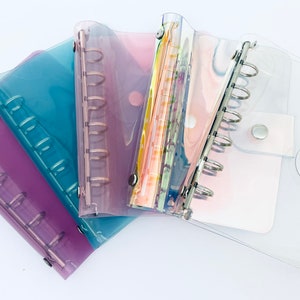 Puede incluir: Un conjunto de cinco carpetas A6 translúcidas en morado, azul verdoso, rosa, iridiscente y transparente. Cada carpeta cuenta con un mecanismo metálico de seis anillas y un cierre de botón a presión. Las cubiertas son de plástico flexible.