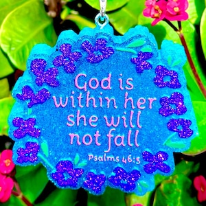 May include: A blue pendant with the text "God is within her she will not fall Psalms 46:5" in pink. The pendant is decorated with purple glitter flowers and has a silver loop for hanging.