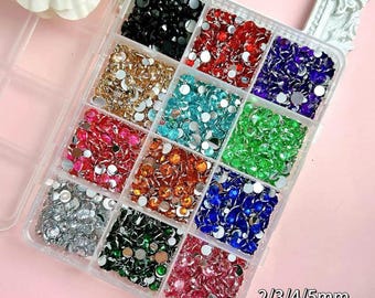 Pode incluir: Um recipiente de armazenamento de plástico transparente com doze compartimentos quadrados preenchidos com pequenos strass redondos e facetados em várias cores, incluindo preto, vermelho, azul, dourado, turquesa, verde, rosa, laranja, prata e roxo, rotulados 2/3/4/5mm.