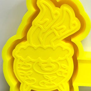 May include: A bright yellow cookie cutter in the shape of a cauldron with flames rising from it. The cauldron has decorative swirls and three small feet. The cutter has a layered design.